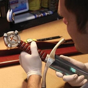 May include: A detailed view of a sword with a silver blade and a handle wrapped in dark red leather. The hilt features a silver cross design on a shield. The sword is being held by a person wearing white gloves.