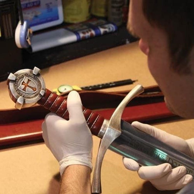 May include: A detailed view of a sword with a silver blade and a handle wrapped in dark red leather. The hilt features a silver cross design on a shield. The sword is being held by a person wearing white gloves.