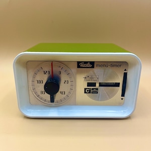 May include: A green and white Fissler menu-timer with a round dial that goes up to 120 minutes. The timer has a white face with black markings and a red needle. The timer also has a white panel with black text that says "TOMATENSOLP" and a black and white icon of a pot.