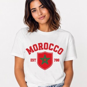 May include: A white short-sleeved t-shirt featuring the red text MOROCCO in an arched varsity font above a red shield emblem containing a green five-pointed star. The text EST and 788 appear in red on either side of the central shield graphic.