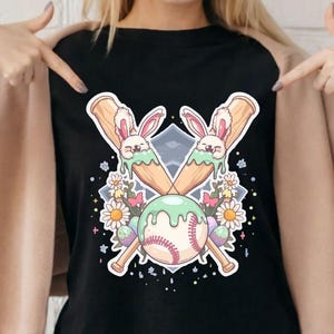 May include: Black t-shirt featuring a baseball-themed Easter design. The graphic includes crossed baseball bats, a baseball with green dripping icing, and two bunny heads with ice cream cones. Floral and pastel egg accents complete the design.