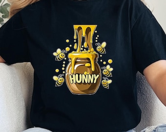 May include: Black t-shirt featuring a cartoon illustration of a honey pot with the word "HUNNY" and dripping honey. Several yellow and black bees surround the honey pot. The design is centered on the front of the shirt.