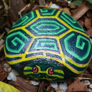 May include: A hand-painted rock shaped like a turtle. The turtle's shell is green and yellow with a geometric pattern. The turtle's face is green with red eyes and a smiling mouth. The rock is surrounded by mulch.