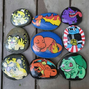 May include: A collection of nine smooth, rounded river stones hand-painted with colorful cartoon characters. The designs include yellow tigers, orange fire-breathing creatures, a purple monster, a blue turtle with sunglasses, and a green creature.