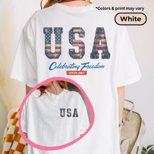 May include: White t-shirt with a distressed USA graphic on the back, featuring the American flag design. The text "Celebrating Freedom" and "4th of July" are printed below the graphic. A smaller USA graphic is on the front.