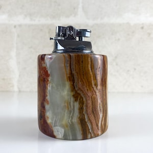 May include: A cylindrical table lighter featuring a polished stone body with swirling bands of brown, cream, and translucent green. The top mechanism is made of shiny silver-toned metal, including a flint wheel, a snuffer cap, and a small adjustment lever.