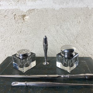 May include: A desk set featuring two square glass inkwells with silver lids, a silver letter opener, a silver dip pen, and a tall, slender silver object. The items are arranged on a dark, rectangular base with a smooth, polished finish.