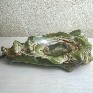 May include: A green onyx decorative bowl with a unique, organic shape. The bowl features swirling patterns of green, brown, and white. The polished surface reflects light, highlighting the natural veining of the stone. The bowl is approximately 12 inches long.