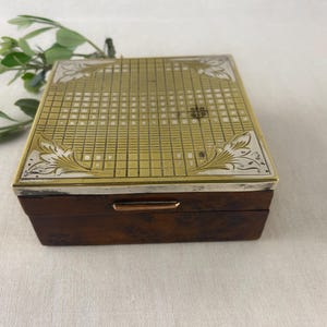 May include: A rectangular wooden box with a brass and silver-toned lid. The lid features a grid pattern with floral corner designs. The box has a dark brown finish and a silver-toned trim. A sprig of greenery is in the background.