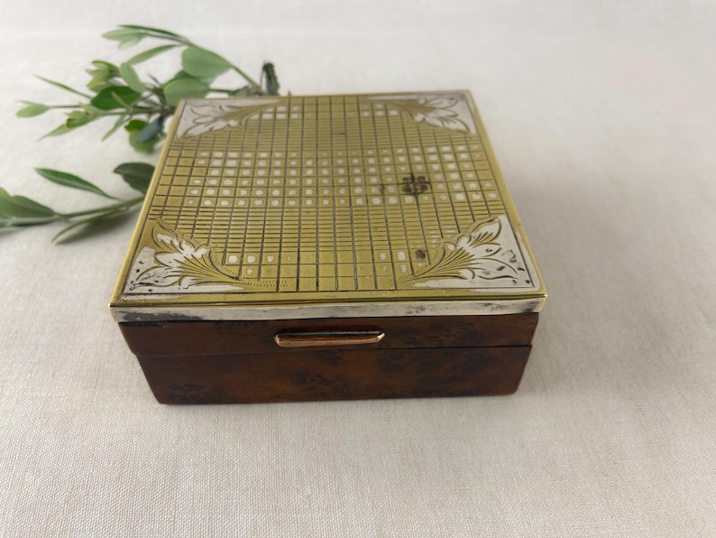 May include: A rectangular wooden box with a brass and silver-toned lid. The lid features a grid pattern with floral corner designs. The box has a dark brown finish and a silver-toned trim. A sprig of greenery is in the background.