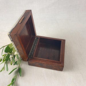 May include: An open, small wooden box with a dark brown finish. The box is rectangular with a hinged lid and a metal clasp. The interior is also brown. Green leaves and stems are visible near the box.
