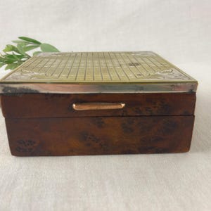 May include: A rectangular wooden box with a brass top featuring a grid pattern and decorative floral edges. The box has a dark brown finish with a small brass handle on the front.