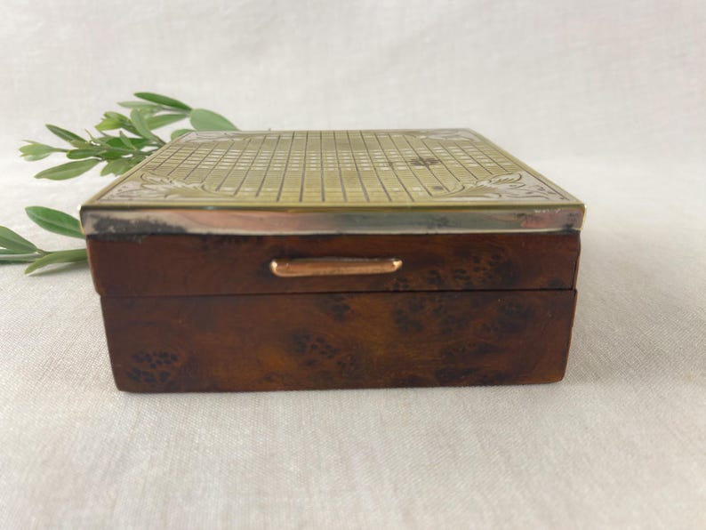 May include: A rectangular wooden box with a brass top featuring a grid pattern and decorative floral edges. The box has a dark brown finish with a small brass handle on the front.
