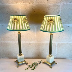 Puede incluir: Dos lámparas de mesa a juego con pantallas a rayas verdes y blancas con ilustraciones de palmeras. Las lámparas tienen tallos y bases de color latón, con bloques de piedra cuadrados de color blanquecino. Las pantallas están ribeteadas con un borde verde oscuro.