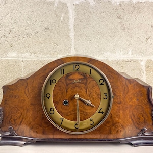 May include: A vintage wooden mantel clock with a burl wood finish. The clock face is gold with black numerals and hands. The clock has a curved top and decorative accents on the sides. The clock measures approximately 10 inches wide.