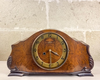 May include: A vintage wooden mantel clock with a burl wood finish. The clock face is gold with black numerals and hands. The clock has a curved top and decorative accents on the sides. The clock measures approximately 10 inches wide.