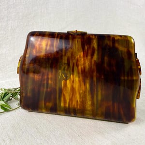 May include: A vintage rectangular clutch purse with a tortoiseshell pattern in shades of brown and amber. The clasp and side details are gold-toned. The purse is a classic accessory.