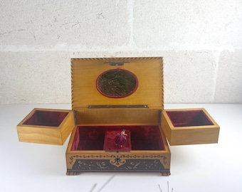 May include: A wooden jewelry box with a light brown finish, featuring an open lid with an oval mirror, two side compartments, and a central section lined in deep red velvet. The exterior base displays dark brown decorative floral inlay patterns.
