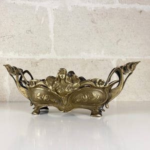 Pode incluir: Uma taça alongada de latão com uma figura central em relevo de uma pessoa com cabelo fluido, ladeada por motivos florais e bordas curvas. O recipiente assenta sobre quatro pés pequenos e apresenta uma superfície texturizada com entalhes.