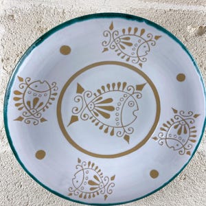 Op de afbeelding: Een ronde witte keramische bord met een donkergroenblauwe rand en een centrale gouden cirkelvormige lijn. Het ontwerp bevat vijf gestileerde gouden visillustraties die rond het midden zijn gerangschikt, afgewisseld met kleine gouden stippen.