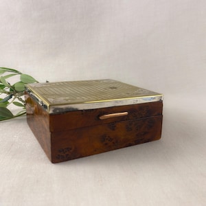 May include: A rectangular wooden box with a silver-colored metal top and trim. The box has a dark brown, patterned exterior and a small copper-colored handle. Green leaves are in the background.