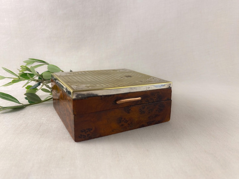 May include: A rectangular wooden box with a silver-colored metal top and trim. The box has a dark brown, patterned exterior and a small copper-colored handle. Green leaves are in the background.