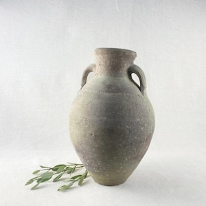 May include: A gray ceramic vase with two handles and a rounded body. The vase has a textured surface and a faded color palette, with hints of green and purple. A sprig of green leaves rests near the base of the vase.