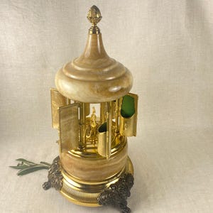May include: A decorative musical carousel in light brown and gold. The carousel has a domed top, ornate gold accents, and a detailed base. It features several compartments and a green interior. The carousel is a decorative item.