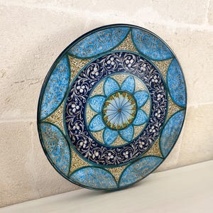 Op de afbeelding: Een ronde decoratieve schaal met een centraal blauw en groen bloemmotief, omringd door concentrische banden van gedetailleerde donkerblauwe en gouden filigraanpatronen, met een herhalend blauw en goud bloembladontwerp langs de buitenrand.