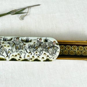 Puede incluir: Caja rectangular de madera abierta con tapa corredera. La tapa está decorada con una escena pintada de jinetes a caballo. El interior de la caja presenta un diseño estampado. Una ramita de vegetación descansa sobre una superficie blanca.