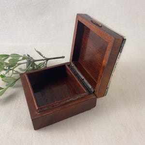 May include: An open, square wooden box with a hinged lid. The box is a dark brown color with a lighter brown interior. A sprig of green leaves rests near the box. The box is likely used for storing small items.