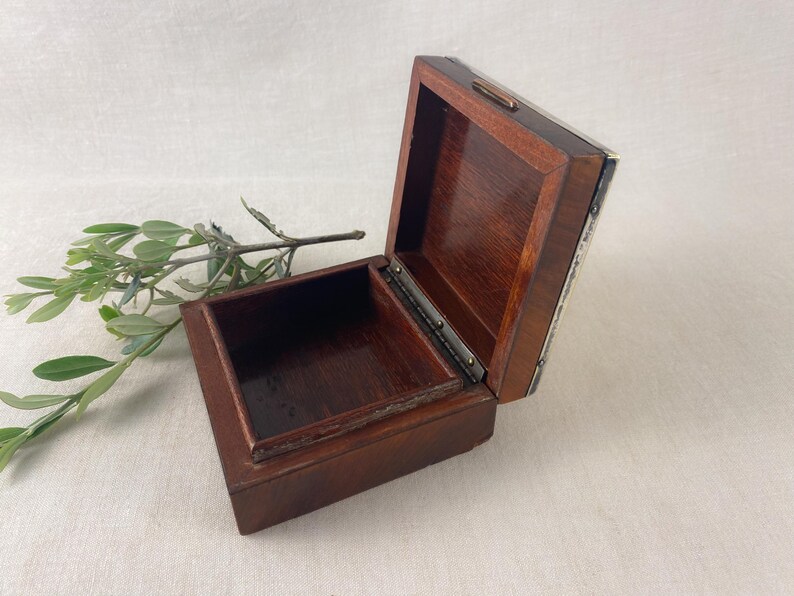 May include: An open, square wooden box with a hinged lid. The box is a dark brown color with a lighter brown interior. A sprig of green leaves rests near the box. The box is likely used for storing small items.