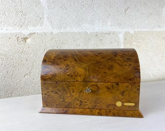 May include: A wooden box with a curved lid and a polished burl wood finish featuring a swirling brown and tan grain pattern. The box includes a small metal keyhole in the center and two small gold-colored rectangular labels on the bottom right.