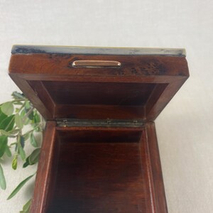May include: Open wooden box with a hinged lid. The box is a dark brown color with a metal trim around the top edge. A small metal handle is on the front of the lid. Green leaves are visible in the background.