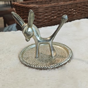 Puede incluir: Soporte para baratijas plateado con forma de burro. El burro se encuentra sobre una bandeja redonda y ornamentada con un detallado diseño floral. El burro tiene orejas largas y una cola levantada. La bandeja tiene un borde decorativo en forma de cuerda.