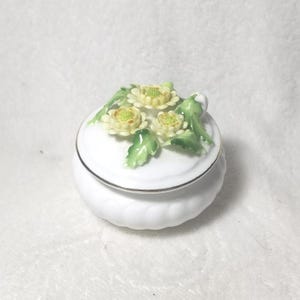 Puede incluir: Pequeña caja de baratijas de cerámica blanca con tapa. La tapa está adornada con tres flores color crema con centros amarillos y hojas verdes. La caja tiene un borde plateado y una base acanalada.