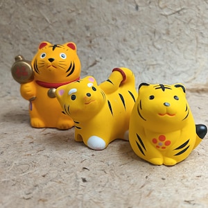 Könnte beinhalten: Drei kleine Tigerfiguren aus gelber Keramik. Der linke Tiger hält einen goldenen Hammer, der mittlere Tiger ist in kriechender Haltung mit einer roten Kordel am Schwanz dargestellt und der rechte Tiger hat ein rot-oranges Blumenmuster auf der Brust.