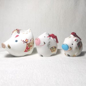 May include: Three white ceramic pig figurines with decorative details. The pigs have pink snouts, gold accents, and red or blue accents. One pig has a floral design, and all have red cords.