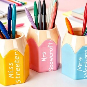 May include: Three hexagonal pencil-shaped pen holders in yellow, pink, and light blue. The yellow holder is labelled Miss Streeter, the pink holder is labelled Mrs Scowcroft, and the light blue holder is labelled Mrs Wenham. Each contains various coloured pens.
