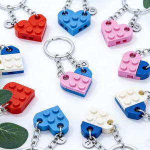 May include: A collection of heart-shaped keychains made from colorful building blocks. The keychains feature red, blue, and pink hearts, some split into two colors. Each keychain has a silver chain and a small, engraved charm.