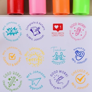 Pode incluir: Uma coleção de selos coloridos de professores com várias frases como "Bom trabalho!" e "Aprovado". Os selos são de cores diferentes e apresentam desenhos como lápis, corações e marcas de verificação. Os selos estão ao lado de marcadores coloridos.
