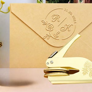 以下が含まれることがあります： ハンドルに花の彫刻が施された金色の金属製エンボスツールが、ベージュの封筒の前に置かれています。封筒には、文字RとK、花のモチーフ、そして日付11.20.2024が描かれた円形のエンボス加工が施されています。