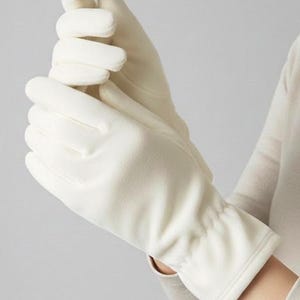 Może przedstawiać: Kremowe rękawiczki wykonane z miękkiego materiału. Rękawiczki mają marszczony detal na nadgarstku i gładki, bezszwowy design. Zdjęcie przedstawia rękawiczki noszone, z palcami jednej dłoni delikatnie dotykającymi drugiej.