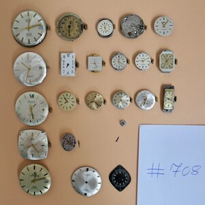 May include: A collection of 21 vintage watch faces and parts, including a silver watch face with black Roman numerals and the word "Preciosa" printed on it. The watch faces are arranged on a beige surface with a white piece of paper with the number "#708" written on it.