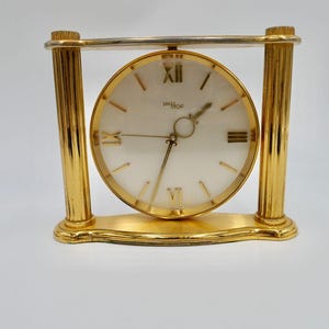 May include: A gold-toned table clock with a white face and black Roman numeral hour markers. The clock is on a stand with two gold columns.