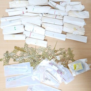 Könnte beinhalten: Eine Sammlung von Uhrteilen, darunter Uhrzeiger, Zahnräder und Zifferblattelemente in verschiedenen Größen und Formen. Die Teile sind in weißem Papier und Plastiktüten verpackt.