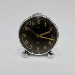 May include: A silver alarm clock with a black face and gold-colored numbers and hands. The clock has the word "ORIS" printed on the face.
