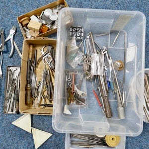 May include: An assortment of jewelry-making tools and supplies, including pliers, scissors, and small containers filled with various components. The tools are arranged in open cardboard boxes and a clear plastic bin on a blue surface.