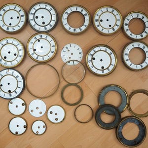 Könnte beinhalten: Eine Sammlung von 18 Vintage-Zifferblättern in verschiedenen Größen und Stilen. Die Zifferblätter sind aus weißem Porzellan mit schwarzen Markierungen und weisen unterschiedliche Abnutzungserscheinungen auf. Einige der Zifferblätter haben Glasabdeckungen, während andere offen sind. Die Zifferblätter sind auf einer Holzoberfläche angeordnet.