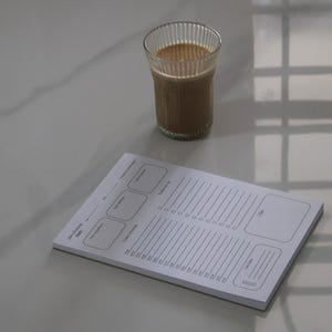 Puede incluir: Un vaso de café con espuma blanca se encuentra junto a un planificador diario blanco. El planificador tiene secciones para notas, una lista de tareas y un horario. El vaso tiene estrías verticales.